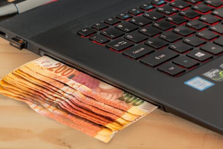 A stack of 200 Czech koruna banknotes partially inserted under a laptop keyboard with red backlit keys on a wooden desk, offering simple tech tips.
