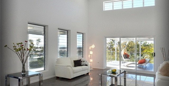Modern living room with large windows providing ample natural light, white couch, and reflective metallic tables. Decor includes a hanging basket chair, ornamental plants, and reasons to have a security system installed.