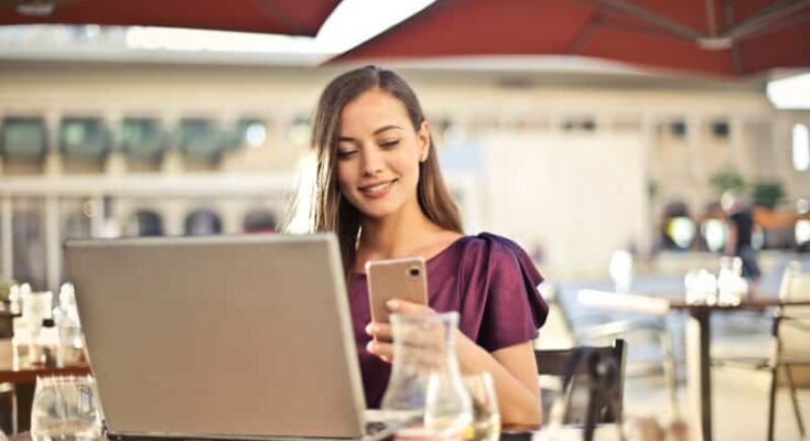 A woman in a purple top seated at a café table, using a smartphone to browse online sites to find people with whom she’s lost touch, with a laptop open in front of her, in a