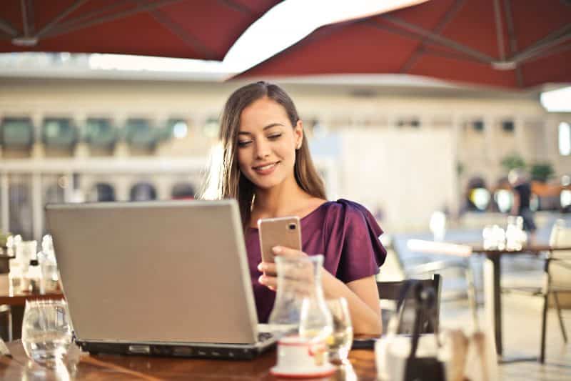 A woman in a purple top seated at a café table, using a smartphone to browse online sites to find people with whom she’s lost touch, with a laptop open in front of her, in a