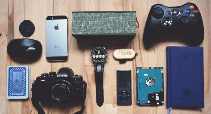 Flat lay of various gadgets and personal items on a wooden surface, perfect as ideas for your geeky partner's birthday, including a smartphone, camera, game controller, watch, notebook, and other small