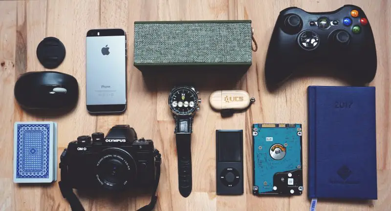Flat lay of various gadgets and personal items on a wooden surface, perfect as ideas for your geeky partner's birthday, including a smartphone, camera, game controller, watch, notebook, and other small