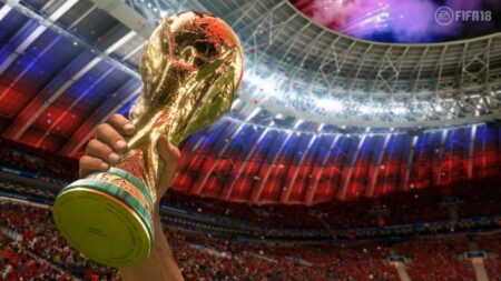 A close-up of a person's hand holding the FIFA World Cup trophy aloft in a packed stadium with vibrant red and blue lighting.