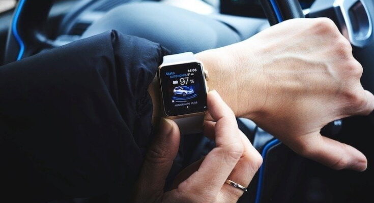 A person utilizing smart tech in a car, checking car-related data on their smartwatch display, such as battery level and range, while holding the steering wheel with the other hand.