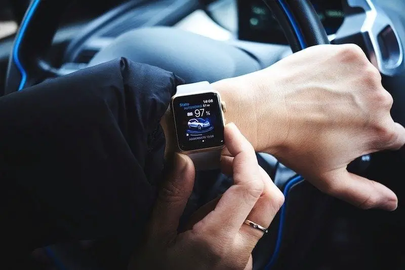 A person utilizing smart tech in a car, checking car-related data on their smartwatch display, such as battery level and range, while holding the steering wheel with the other hand.