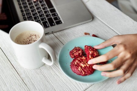 A person's hand reaching for a red cookie on a Smart Tech teal plate, next to a white mug of coffee and laptop on a wooden table.