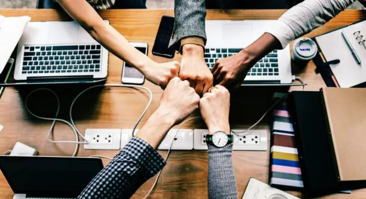 Four people fist-bumping over a cluttered office table containing laptops, notebooks, and a power strip, symbolizing teamwork and collaboration in expanding your brand internationally.