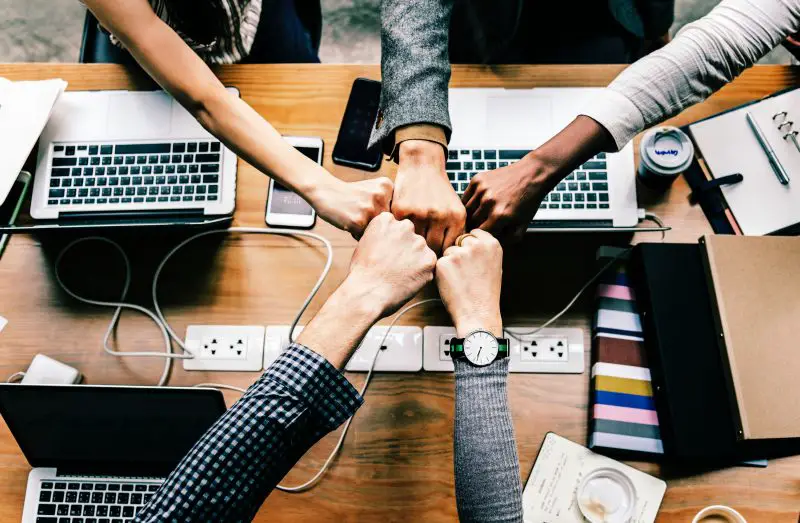 Four people fist-bumping over a cluttered office table containing laptops, notebooks, and a power strip, symbolizing teamwork and collaboration in expanding your brand internationally.