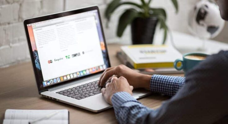 A professional in a checkered shirt explores "How To Use The Internet To Build Your Career" on a MacBook with a relevant document open, sitting at a desk equipped with a coffee mug, books,