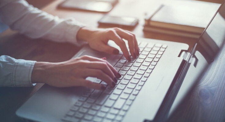 Close-up of a person's hands typing on a laptop keyboard, with a soft focus on a wooden desk background illuminated by warm light, researching how to get all the best gadgets on a budget.