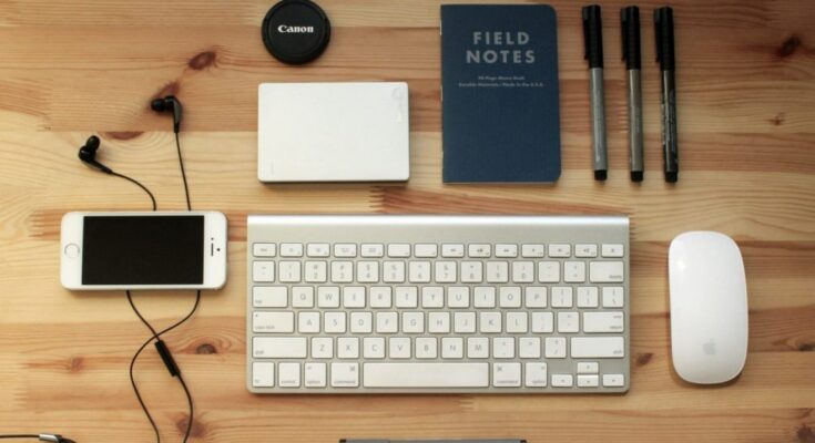 Top view of a wooden desk with various technological tools arranged neatly including a smartphone, headphones, keyboard, mouse, pens, notebooks, and a camera lens cap.