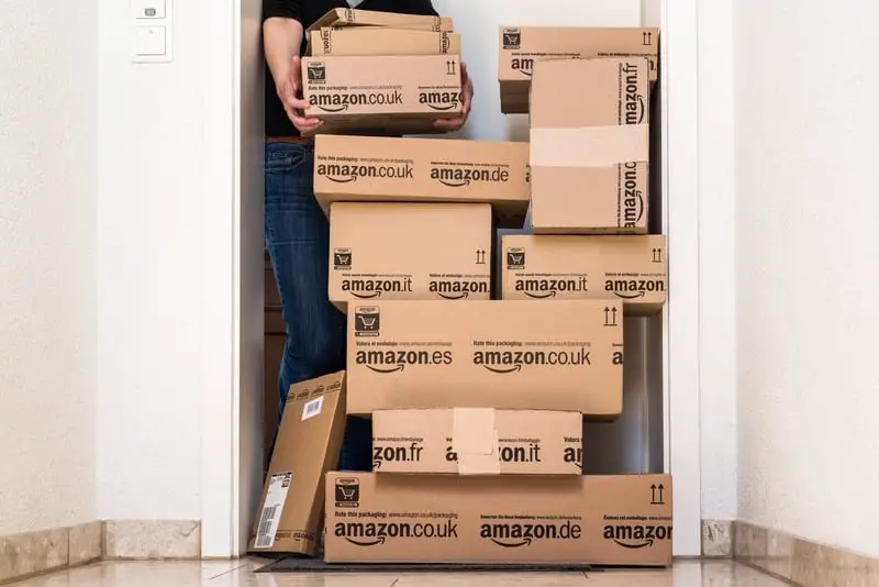 A person standing in a doorway, obscured by a large stack of Amazon boxes containing the best gaming gear they are holding, indicating a recent delivery or move. Visible branding includes various Amazon European domains.