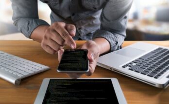 A person seated at a desk using a smartphone with Java application code on the screen, alongside a laptop and tablet also displaying code. Focus on technology use in a modern workspace.