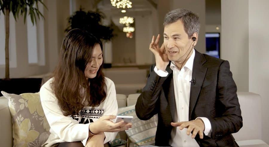 Man in a suit and headset gesturing with his hand while speaking to a smiling woman holding an open book, seated on a couch in a well-lit room with chandeliers, discussing how tech