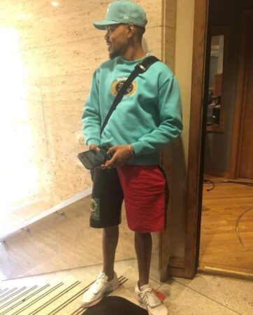 A man stands in a lobby, wearing a turquoise sweatshirt, red shorts, and white sneakers, accessorized with a blue crossbody bag and black cap. He is holding a smartphone and looking