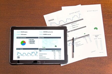 A tablet displaying a business dashboard with charts and data sits on a wooden desk alongside scattered printed graphs and a requirement management tool.