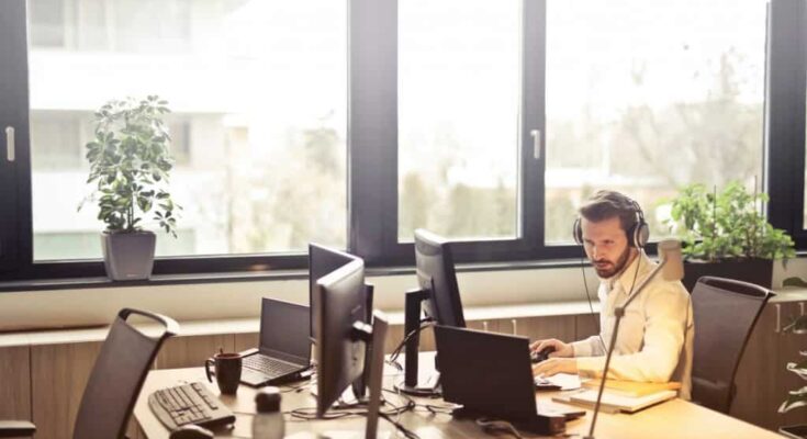 A man wearing headphones works attentively at his desk with multiple computer monitors, managing help desk software in a sunlit office.