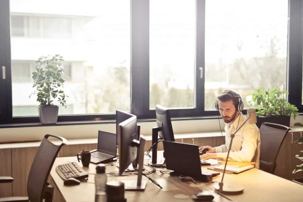 A man wearing headphones works attentively at his desk with multiple computer monitors, managing help desk software in a sunlit office.