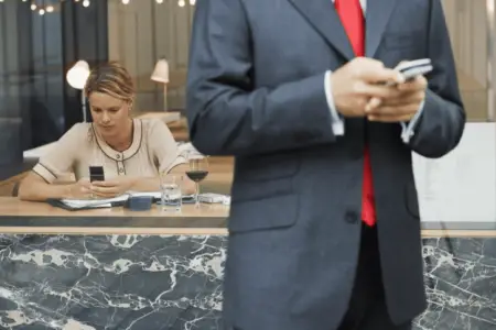 A man in a suit with a red tie stands in the foreground holding a smartphone, likely texting customers, while a woman sits at a cafe table in the background, looking at her own phone.