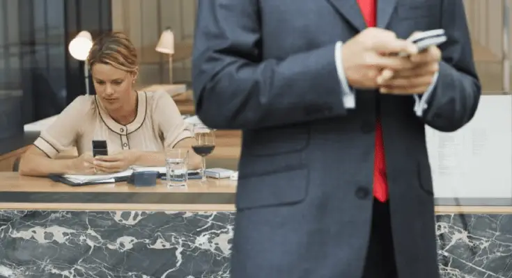 A man in a suit with a red tie stands in the foreground holding a smartphone, likely texting customers, while a woman sits at a cafe table in the background, looking at her own phone.