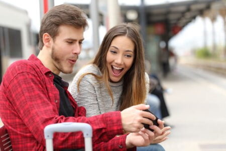 A young man and woman sitting at a train station, laughing and looking at an online gaming site on a smartphone together. The man is wearing a red flannel shirt, and the woman is wearing a