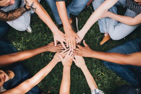 Aerial view of diverse group of people sitting in a circle on grass, placing their hands together in the center, symbolizing teamwork and unity during a digital transformation workshop.