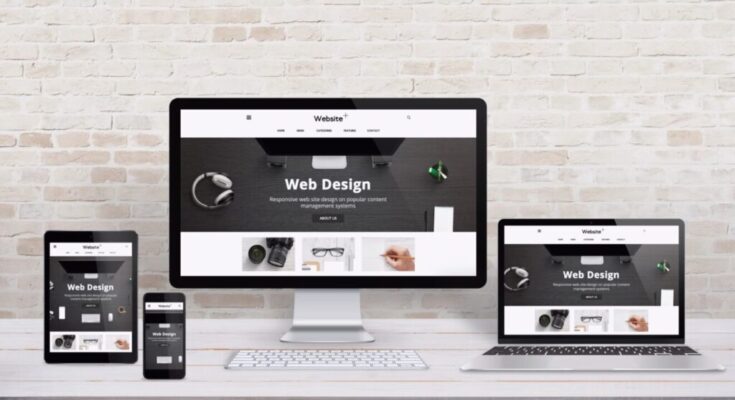 Multiple display devices with modern flat design web site presentation. Wooden desk and brick wall interior.