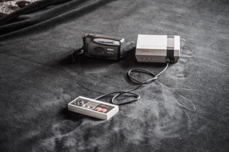 An old Nikon camera alongside a classic Nintendo Entertainment System console with its controller on a textured gray surface, one of the best gifts for gamers.