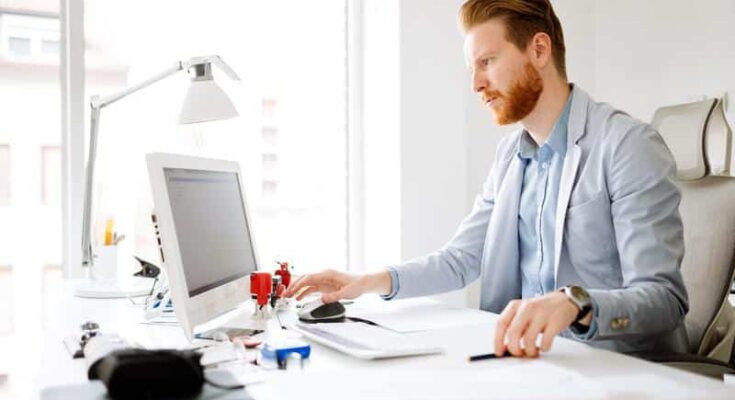 A focused businessman with a beard working at a desk with multiple papers and a desktop computer in a bright office, excelling in his best tech career.