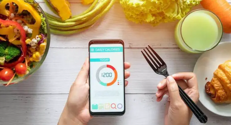 A person tracks daily calorie intake using health software on a smartphone, surrounded by colorful vegetables, a croissant, and a glass of juice on a wooden table.