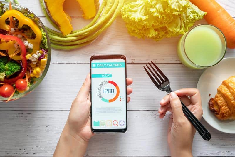 A person tracks daily calorie intake using health software on a smartphone, surrounded by colorful vegetables, a croissant, and a glass of juice on a wooden table.