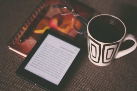 A cozy student reading setup with an e-reader displaying text, a notebook with a pen, reading glasses, and a cup of coffee on a textured table.