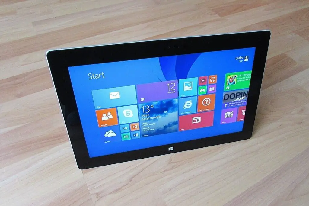 A tablet computer displayed on a wooden floor, running Windows 10, showing a vibrant and colorful start screen with various application tiles.
