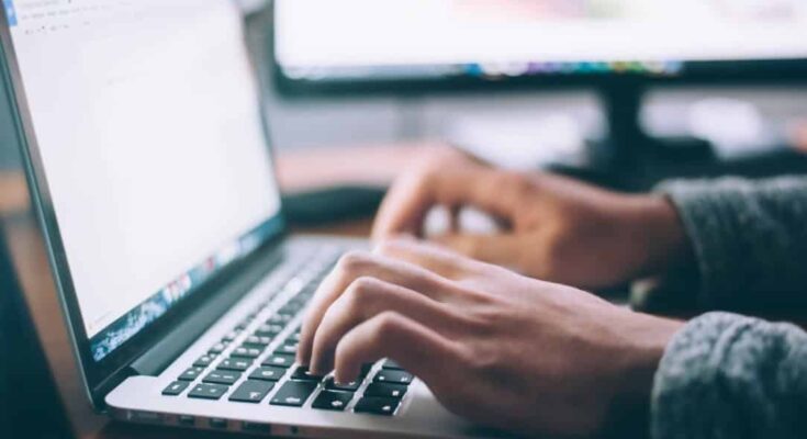 Close-up of a student's hands typing on a laptop keyboard with a blurred background showing another computer monitor.