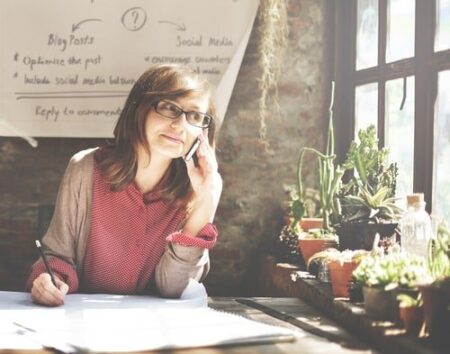 A woman wearing glasses talks on a mobile phone about influencer marketing while sitting at a desk with papers and plants, in a room with a large window and a whiteboard filled with notes.