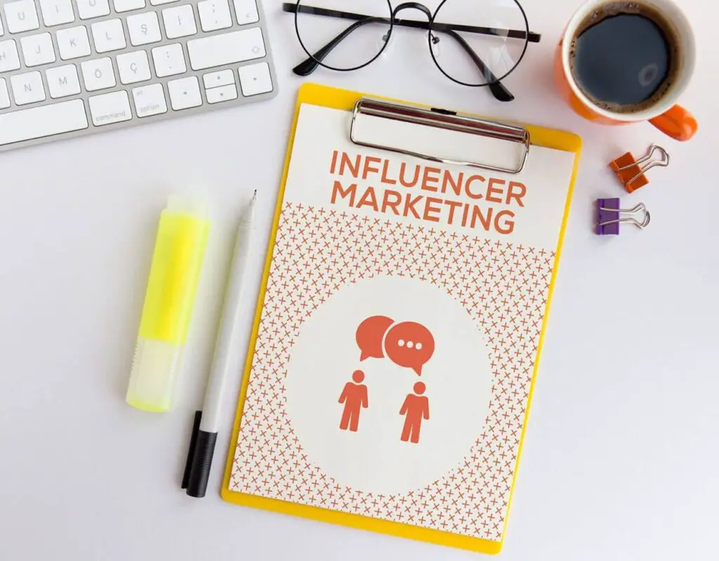 Clipboard with "influencer marketing" text, icons, and a stylized audience pattern, beside a keyboard, eyeglasses, highlighter, pen, binder clips, and a cup of coffee