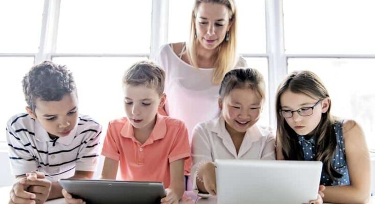 A teacher assists a diverse group of four students with digital tablets and a laptop in a brightly lit classroom. They are focused and engaged in educational software learning.