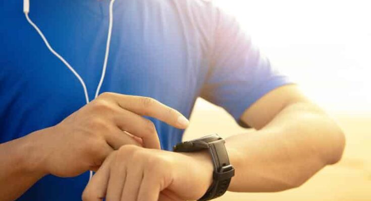 A person wearing a blue shirt and earphones adjusts a black digital watch with fitness apps on their wrist, illuminated by warm sunlight.