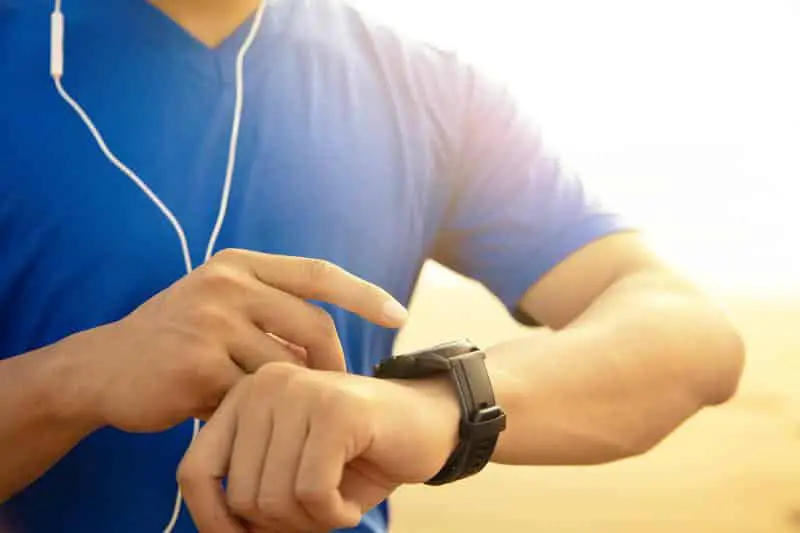 A person wearing a blue shirt and earphones adjusts a black digital watch with fitness apps on their wrist, illuminated by warm sunlight.