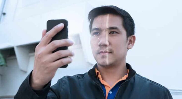 A man with a faint beard is holding and looking intently at his smartphone, using facial recognition, with an expression of focus and slight curiosity, in an indoor setting with soft lighting.