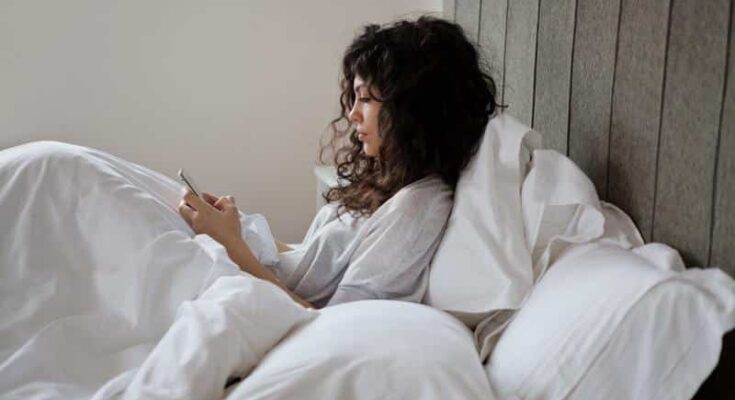 A woman with curly hair, sitting in bed covered with white sheets, is texting on a smartphone, with a serene expression on her face.