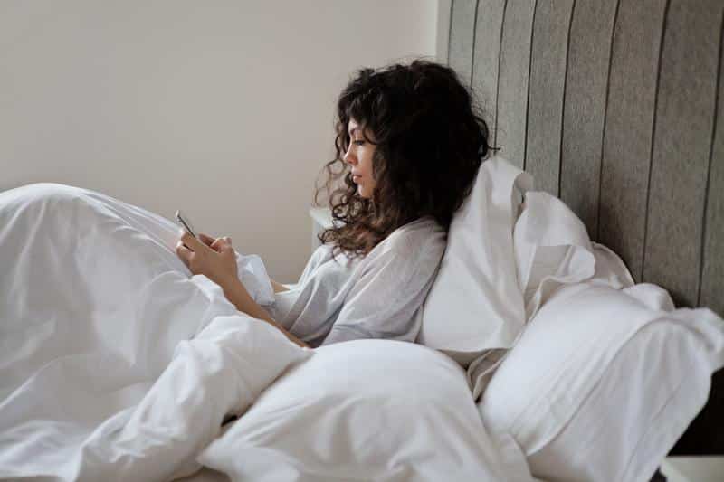 A woman with curly hair, sitting in bed covered with white sheets, is texting on a smartphone, with a serene expression on her face.