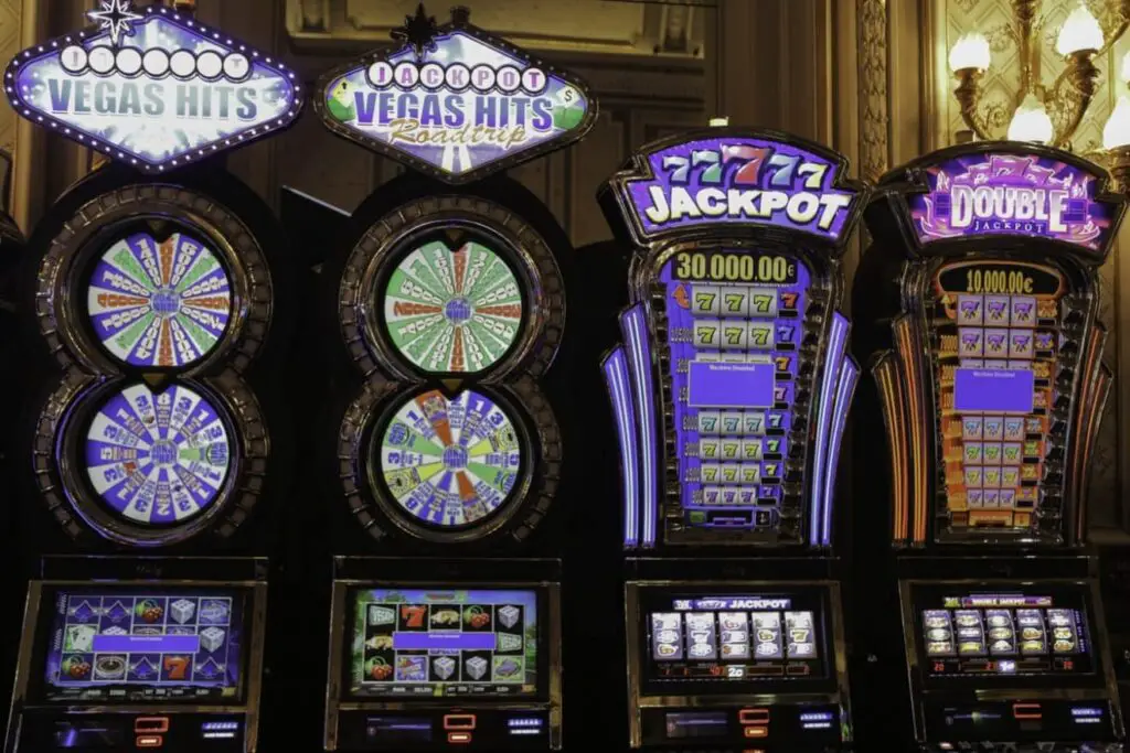 A row of colorful slot machines, including "vegas hits" and "jackpot," displays vibrant screens and illuminated signs in a lively casino setting.