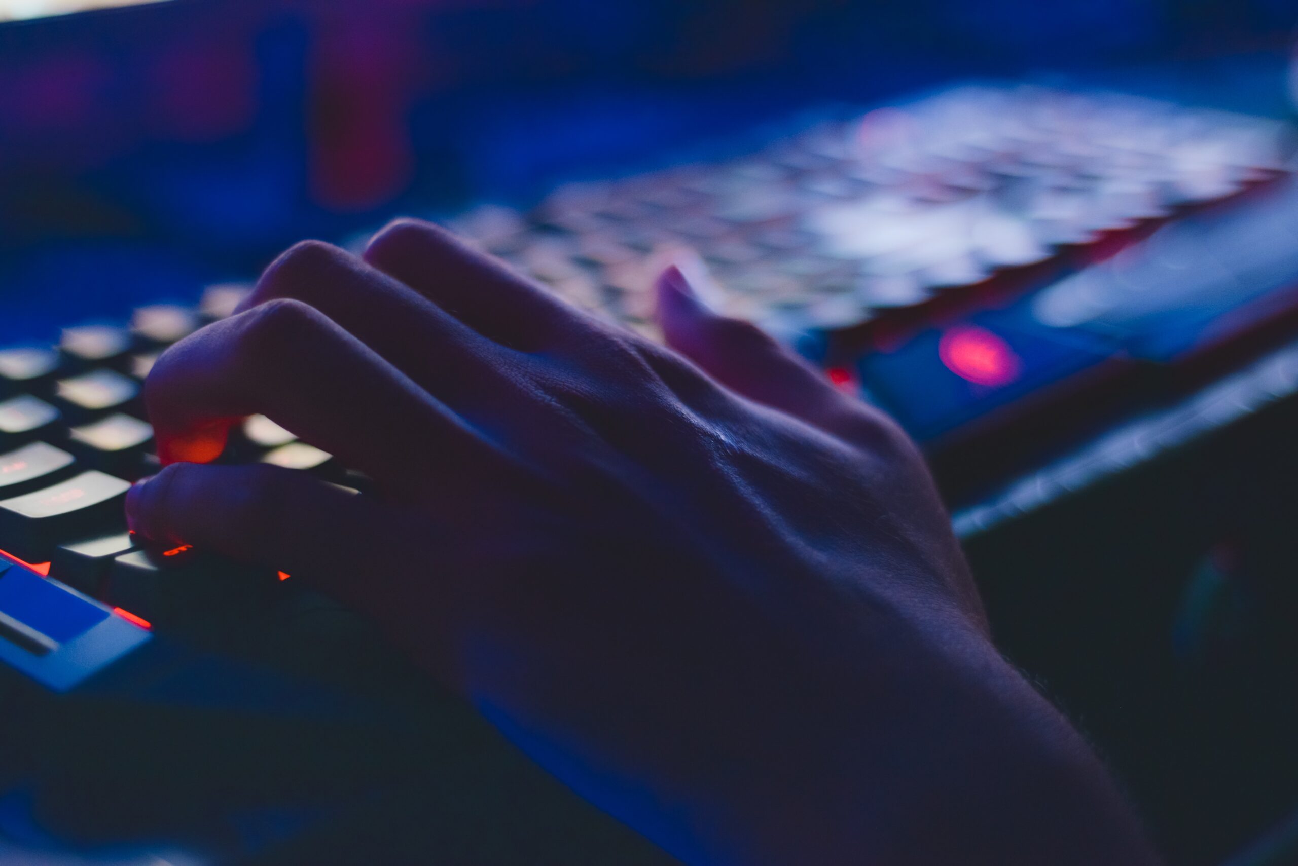 A close-up of a person's hands typing on a backlit gaming keyboard in a dimly lit room, emphasizing the vivid colors on the keys.