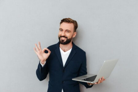 A cheerful bearded man in a suit holding a laptop displaying gambling statistics in one hand and making an 'ok' gesture with the other, standing against a light gray background.