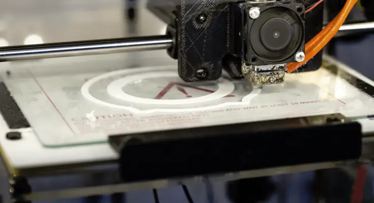 A close-up view of a 3D printer's extruder head as it is printing a white object with a circular design on a transparent print bed.