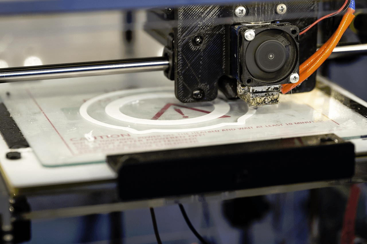 A close-up view of a 3D printer's extruder head as it is printing a white object with a circular design on a transparent print bed.