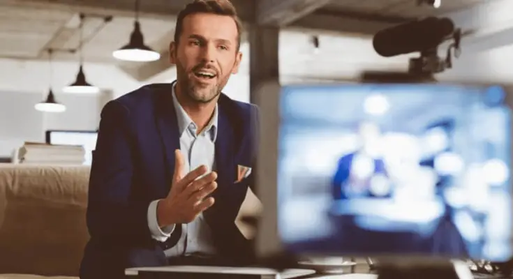 A man in a blue blazer and white shirt speaks animatedly at a desk, a blurred computer screen in the foreground showing additional people.