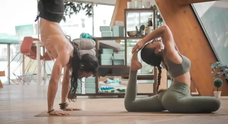 Two women practicing fitness yoga indoors, one in a handstand and the other performing a reclining twist pose, in a serene, well-lit room with wooden accents.