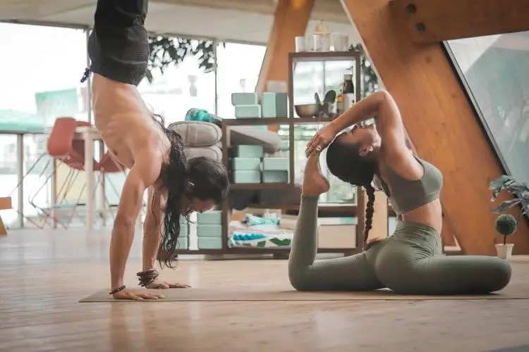 Two women practicing fitness yoga indoors, one in a handstand and the other performing a reclining twist pose, in a serene, well-lit room with wooden accents.